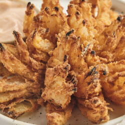 A golden-brown blooming onion served on a white plate with a creamy dipping sauce.