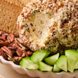 A pecan-crusted cheese ball with a scoop taken out, served with crackers and cucumber slices.