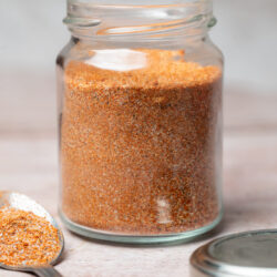 Hamburger seasoning in a glass jar with a spoon of seasoning next to it.