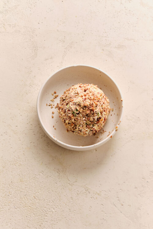 A cheese ball coated in chopped pecans, served in a white bowl.