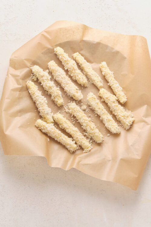 Breaded mozzarella sticks on parchment paper, ready for freezing.