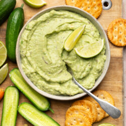 Avocado dip in a bowl, garnished with lime, served with crackers, cucumbers, and carrots.