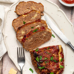 Slices of cheesy meatloaf on a white platter, topped with ketchup glaze and fresh parsley.
