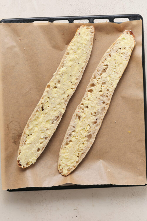 Spreading garlic butter mixture on both halves of French bread for cheesy garlic bread.