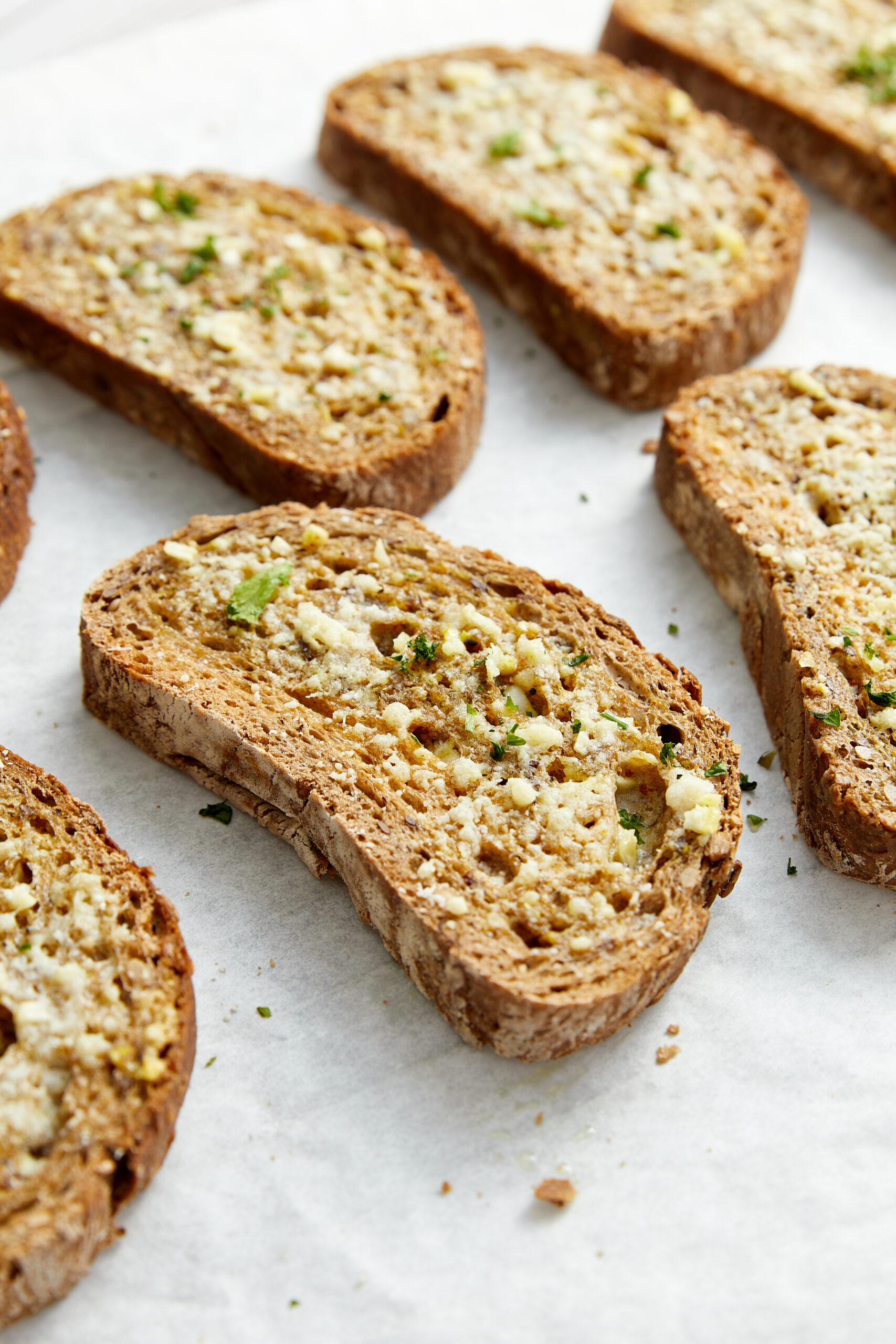 Garlic toast slices on parchment paper, topped with garlic, butter, Parmesan, and fresh parsley.