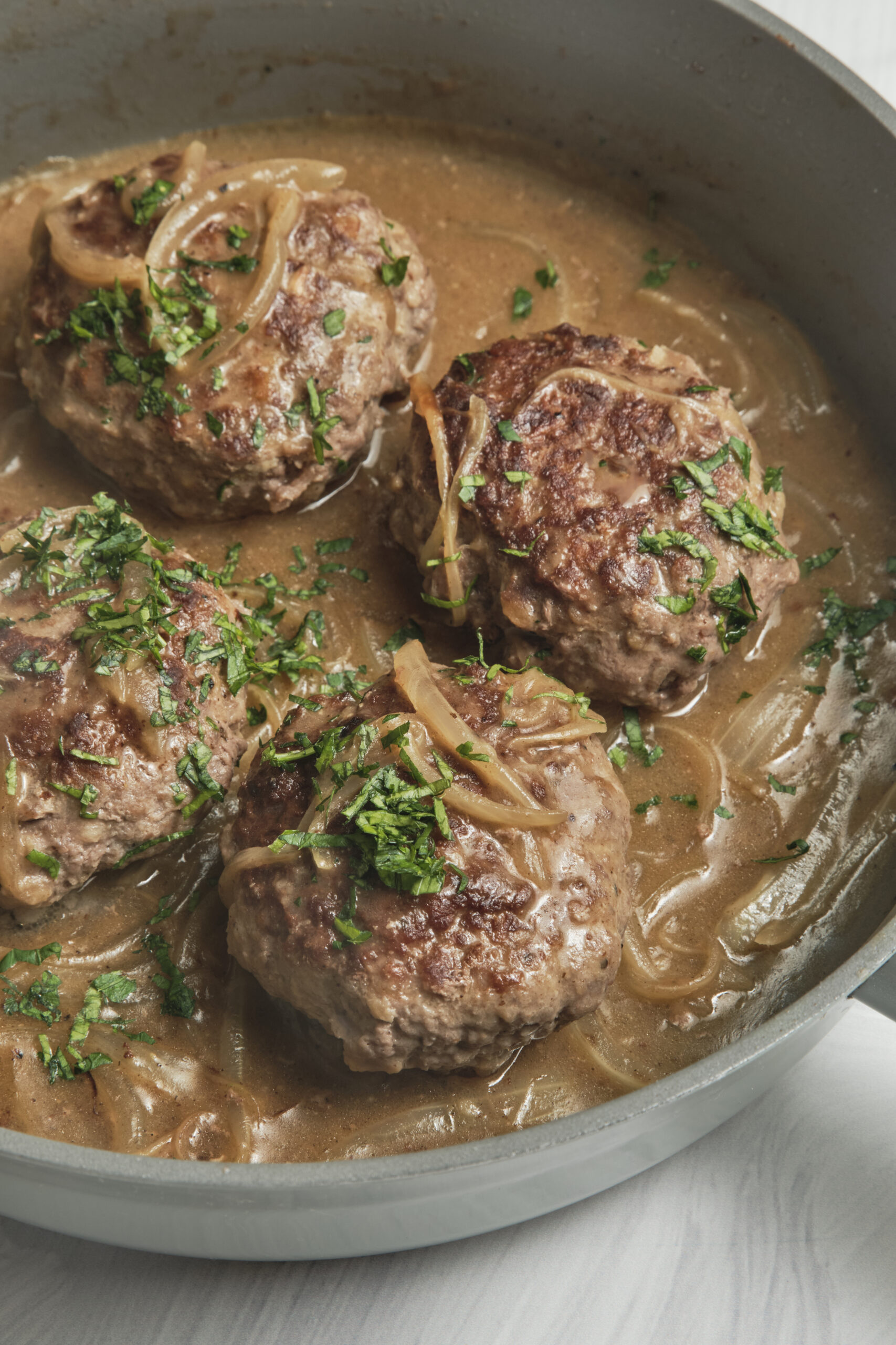 Four hamburger steak patties simmering in a rich brown gravy with sliced onions and fresh parsley.
