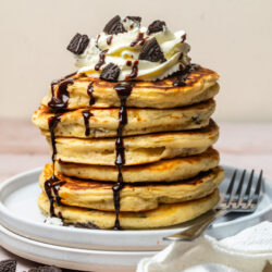 A stack of Oreo pancakes topped with whipped cream, chocolate syrup, and crushed cookies.